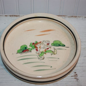 May include: Cream ceramic plate with a black and brown rim. The plate has a hand-painted scene with figures, green bushes, and orange lines. The design is reminiscent of folk art.