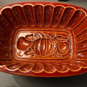 May include: A rectangular, glazed ceramic baking dish in a rich, reddish-brown hue. The dish features a decorative embossed design of a pumpkin and gourds in the center, with a scalloped edge and vertical ridges around the perimeter. A kitchenware item.