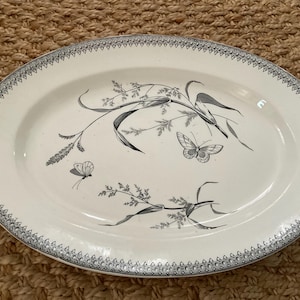 May include: Oval serving platter with a white background and a decorative border. The platter features a black and gray botanical design with butterflies and foliage. The border has a repeating pattern.
