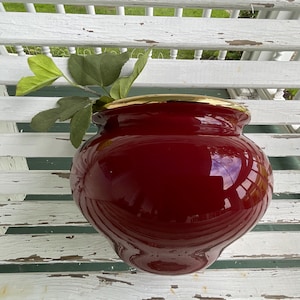 Peut inclure: Un vase en céramique rouge foncé brillant avec un corps arrondi et bulbeux et une surface lisse et réfléchissante. Le vase présente un bord doré brillant distinct autour de l'ouverture supérieure et contient une petite tige de plante verte.
