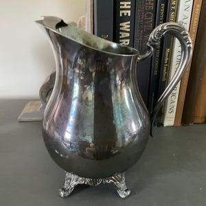 Puede incluir: Una jarra plateada con un asa curva y un pico. La jarra tiene un cuerpo bulboso y descansa sobre tres pies decorativos. El fondo incluye una colección de libros.