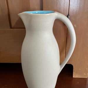 May include: A beige ceramic pitcher with a curved handle and a light blue interior. The pitcher has a slender, elongated shape and a spout for easy pouring. The handle is arched, providing a comfortable grip. The pitcher is likely used for serving beverages.