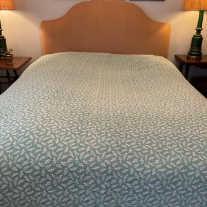 May include: A bed with a light green and white patterned bedspread. The headboard is a light tan color. Two lamps with orange shades sit on either side of the bed. Artwork hangs above the headboard.