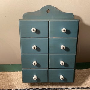 May include: A blue wooden spice rack with eight drawers and white knobs. The rack is mounted on a wall.