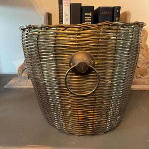May include: A woven metal basket with a handle. The basket is a muted gold color and has a circular handle attached to the front. Several books are visible inside the basket.