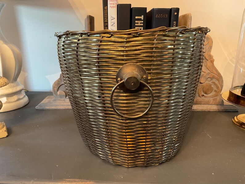May include: A woven metal basket with a handle. The basket is a muted gold color and has a circular handle attached to the front. Several books are visible inside the basket.