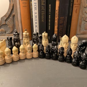 以下が含まれることがあります： 黒とクリーム色の駒が揃ったチェスセット。駒には、ナイト、ルーク、ビショップ、キング、ポーンが含まれます。チェスの駒は、背景に本がある灰色の表面に配置されています。