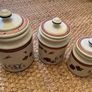 May include: Set of three ceramic canisters with lids. The canisters are white with a pink and red band around the top and bottom. Floral and leaf designs are painted on the sides. The canisters are different sizes.
