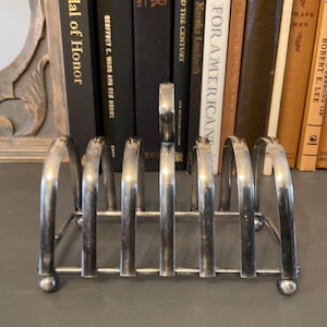 May include: A silver-colored metal toast rack with arched dividers. The rack has a rectangular base and rounded feet. Books are visible in the background.