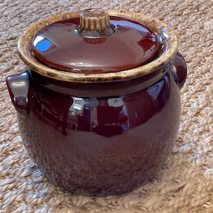 May include: A maroon ceramic crock pot with a matching lid. The pot has a textured exterior and two side handles. The rim and lid edge are a lighter brown color. The lid has a small, rounded handle. This kitchenware is suitable for cooking and serving.