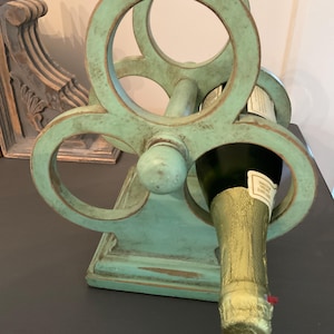 May include: A teal-colored wooden wine bottle holder with a distressed finish. The holder has a circular design and is holding a champagne bottle. The holder is on a dark surface.