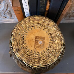May include: A round, woven brass basket with a flat, brass-colored base. The basket has a handle on one side and is sitting on a dark surface. The basket is likely used for storage or decoration.