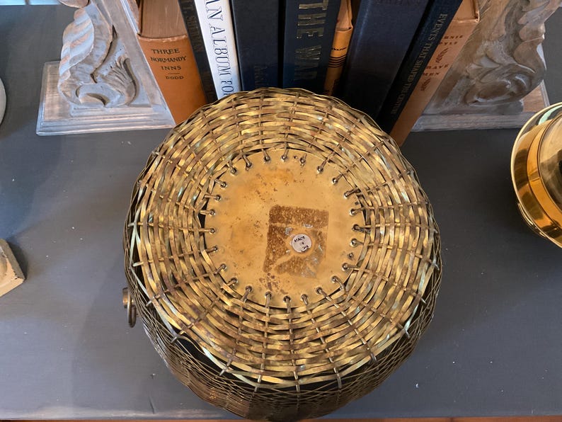 May include: A round, woven brass basket with a flat, brass-colored base. The basket has a handle on one side and is sitting on a dark surface. The basket is likely used for storage or decoration.
