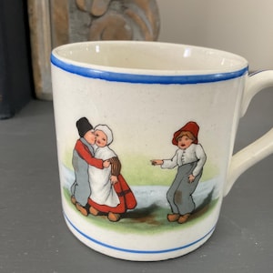 May include: White ceramic mug with a blue rim and a hand-painted scene of three children. Two children are embracing, and the third child is pointing to the side. The mug has a handle.