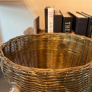 May include: A woven gold-colored basket with a round shape. The basket is made of tightly woven material and has a small, round metal plate attached. Books are visible in the background.