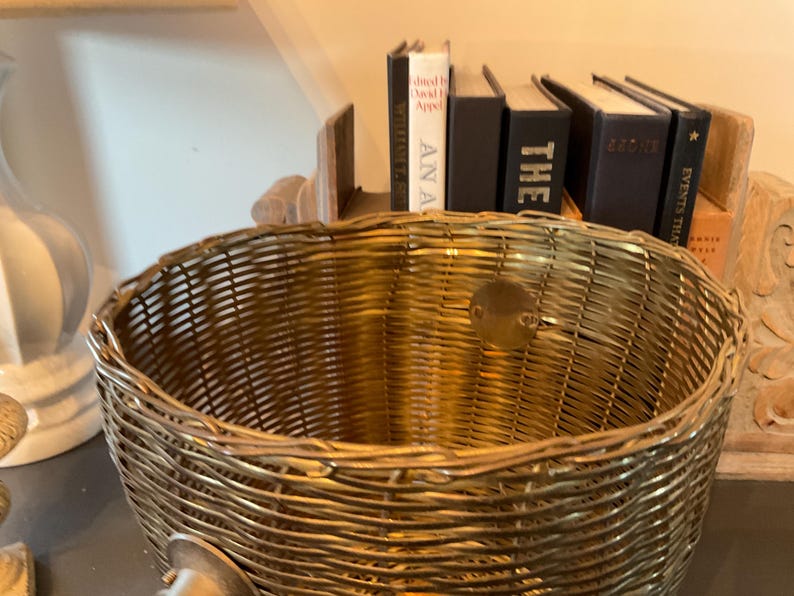 May include: A woven gold-colored basket with a round shape. The basket is made of tightly woven material and has a small, round metal plate attached. Books are visible in the background.