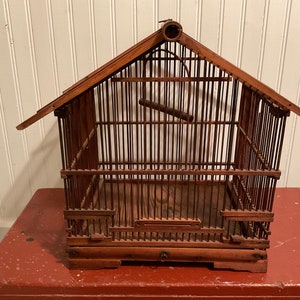 Puede incluir: Una jaula de pájaros de madera vintage con un diseño de techo inclinado. La jaula está construida con barras verticales de madera y presenta una percha en el interior. La jaula es de madera marrón oscuro y se encuentra sobre una superficie roja.