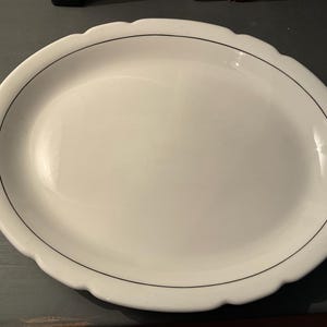 May include: Oval white serving platter with a scalloped edge and a thin black line detail. The platter is empty and appears to be made of ceramic or porcelain. Ideal for serving appetizers or entrees.