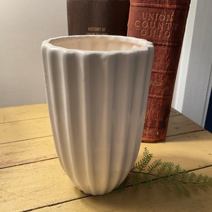 Może przedstawiać: Wysoki, biały ceramiczny wazon z żebrowanym wzorem zewnętrznym. Wazon jest cylindryczny z lekko rozszerzoną górą. Jest umieszczony na żółtej drewnianej powierzchni, z antycznymi książkami w tle.