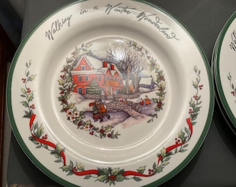 May include: White ceramic plate with a green rim and a winter scene illustration. The plate features a red house, a bridge, and people walking in the snow. The words "Walking in a Winter Wonderland" are written on the plate.