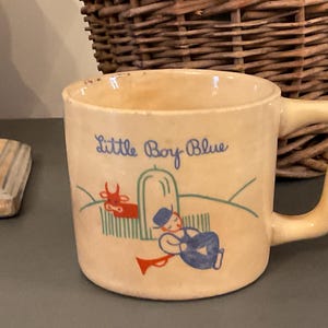 Puede incluir: Una taza de cerámica de color crema con un asa azul. La taza tiene una ilustración colorida de un niño durmiendo junto a una vaca roja. El texto "Little Boy Blue" está impreso en azul en la taza.