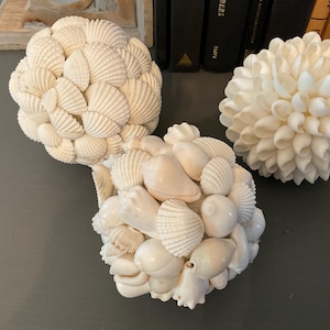 May include: Three spherical decorative objects covered in various white seashells. One sphere features fan-shaped shells, another displays smooth, rounded shells, and the third is composed of pointed, petal-like white shells arranged in a dense cluster.