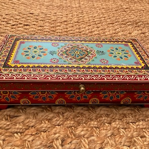 May include: A rectangular wooden box with a red exterior and a decorative, colorful top. The top features a light blue background with floral patterns in orange, yellow, and blue. The box has a brass-colored knob.