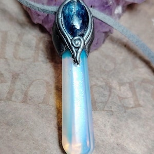 May include: A crystal pendant necklace with a pearlescent crystal point. The crystal is set in a silver-toned metal with a blue stone. The necklace has a grey suede cord.