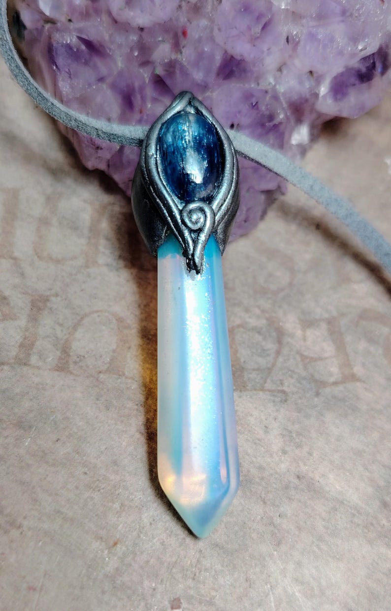 May include: A crystal pendant necklace with a pearlescent crystal point. The crystal is set in a silver-toned metal with a blue stone. The necklace has a grey suede cord.