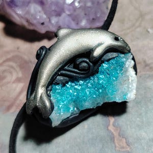 May include: A silver dolphin charm pendant with a blue crystal geode. The dolphin is leaping out of the geode, which is set in black polymer clay.