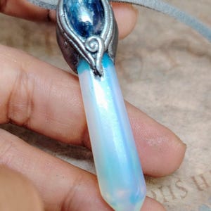 May include: A handcrafted pendant necklace featuring a large, iridescent crystal with a blue gemstone accent. The crystal is a pale blue and white colour, and the pendant is attached to a grey suede cord.