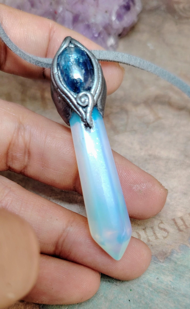 May include: A handcrafted pendant necklace featuring a large, iridescent crystal with a blue gemstone accent. The crystal is a pale blue and white colour, and the pendant is attached to a grey suede cord.