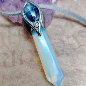 May include: A pendant necklace featuring a light blue crystal with a pearlescent sheen. The crystal is set in a silver-coloured metal casing with a blue gemstone at the top. The necklace has a grey suede cord.