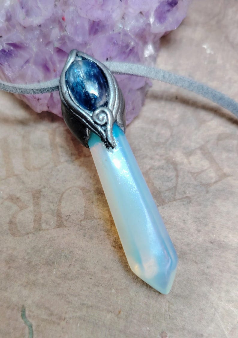 May include: A pendant necklace featuring a light blue crystal with a pearlescent sheen. The crystal is set in a silver-coloured metal casing with a blue gemstone at the top. The necklace has a grey suede cord.