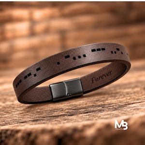 Pode incluir: Uma pulseira de couro castanho escuro com marcas de código Morse a preto na superfície exterior. O interior da tira de couro está gravado com a palavra Forever. A pulseira é fixada com um fecho magnético de metal preto mate.