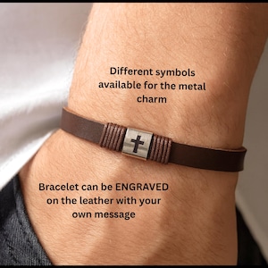 May include: A brown leather bracelet featuring a silver cross charm. The bracelet has a braided design on either side of the charm. The image text indicates that various symbols are available and the bracelet can be engraved.