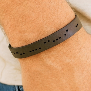May include: Dark brown leather bracelet with Morse code detailing. The bracelet is a simple band with rectangular cutouts forming the code. The bracelet is worn on a wrist.