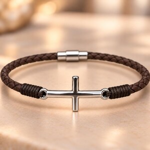 May include: A brown braided leather bracelet featuring a polished silver-tone metal cross centerpiece. The cross is connected to the leather band by dark brown cord wrappings on each side, and the bracelet is secured with a silver-tone magnetic clasp.