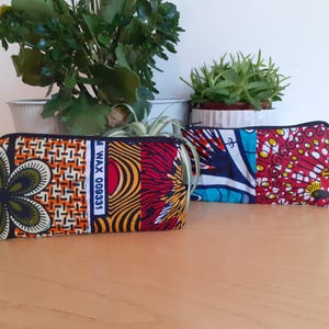 May include: Two colourful fabric zippered pouches with different patterns. The pouch on the left has a black flower, orange, yellow, and red stripes, and the text "M WAX 008331". The pouch on the right has a blue, white, and red pattern.
