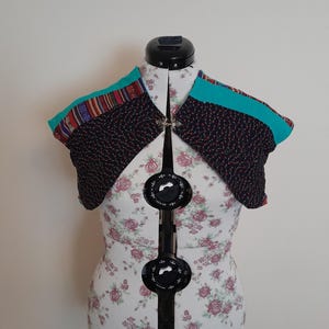 May include: A black quilted bolero with turquoise and multi-coloured patterned fabric trim. The bolero is displayed on a white dress form with floral print.