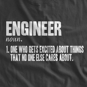 May include: A dark grey t-shirt with white text that reads "ENGINEER noun. 1. ONE WHO GETS EXCITED ABOUT THINGS THAT NO ONE ELSE CARES ABOUT."