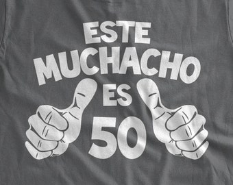 May include: A dark gray t-shirt with white text that reads "ESTE MUCHACHO ES 50" with two thumbs up on either side of the number 50.