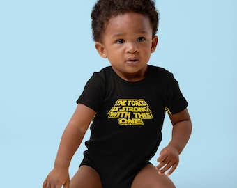 May include: A black baby bodysuit with the text "THE FORCE IS STRONG WITH THIS ONE" in yellow. The baby is sitting on a light blue background, with a yellow star-shaped pillow in the lower left corner.