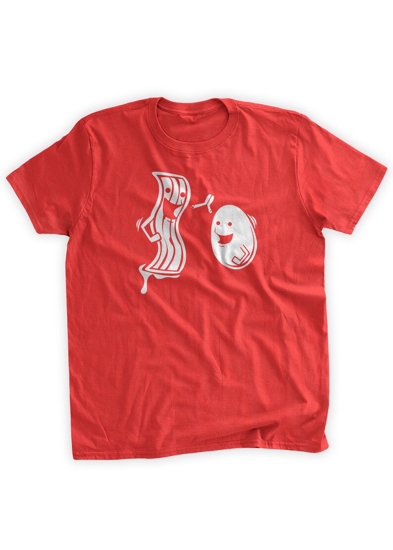 Puede incluir: Camiseta roja con un gr&aacute;fico blanco de una tira de tocino de dibujos animados persiguiendo un huevo de dibujos animados. El tocino tiene una cara y sostiene una esp&aacute;tula. El huevo tiene una cara y sonr&iacute;e.