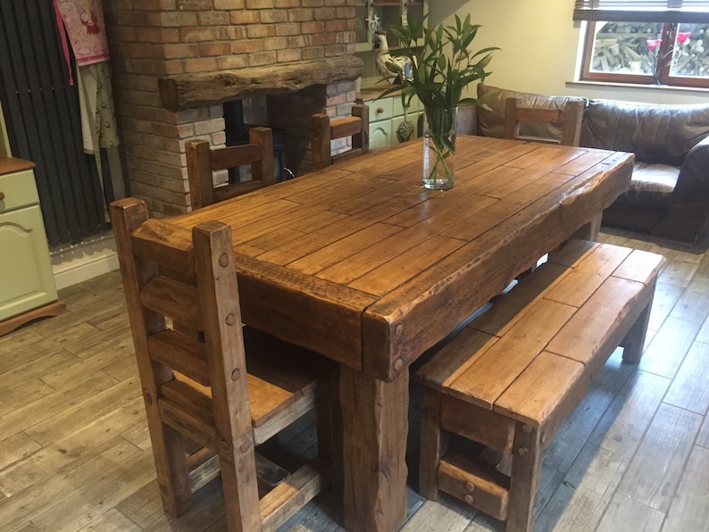 May include: A rustic wooden dining table with matching chairs and a bench. The table is rectangular, with a natural wood finish and visible grain. A clear glass vase with green stems sits on the table. The chairs and bench have a similar design.