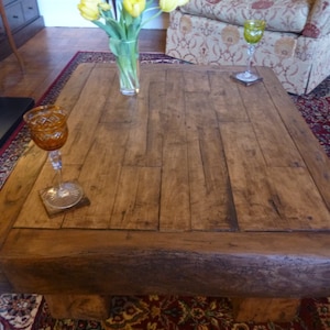 May include: A rustic wooden coffee table with a thick, dark brown wood top and a natural wood finish. The table has a rectangular shape and sits on four sturdy legs. The table is in a living room setting with a rug and a vase of yellow tulips.