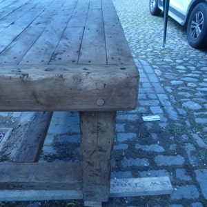 May include: A large, rustic wooden table with a thick, weathered top and sturdy legs. The table is made of reclaimed wood and has a natural, distressed finish.