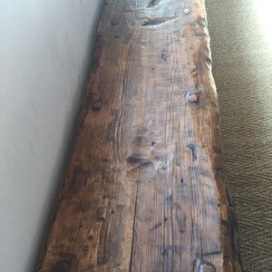 May include: A long, rustic wooden bench with a natural, dark brown finish. The bench has a live edge and is made from reclaimed wood. The wood has a lot of character and shows signs of age.