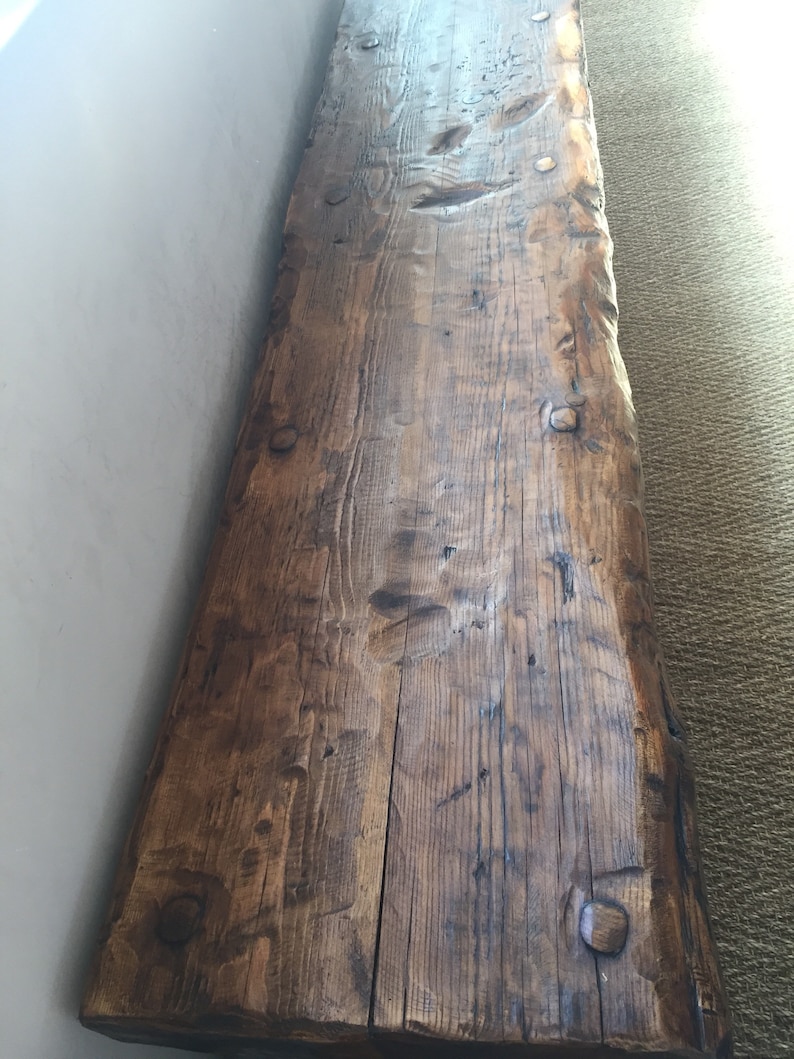 May include: A long, rustic wooden bench with a natural, dark brown finish. The bench has a live edge and is made from reclaimed wood. The wood has a lot of character and shows signs of age.