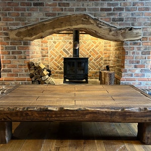 May include: A large, rustic wooden coffee table with a thick, dark brown top and sturdy legs. The table is in front of a brick fireplace with a wood-burning stove.